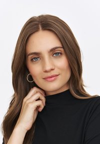 Portret osoby z długimi brązowymi włosami, ubranej w czarną bluzkę z wysokim kołnierzem. Złote kolczyki w kształcie obręczy i delikatny makijaż podkreślają jej rysy.