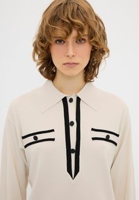 Camicia a maniche lunghe crema con patta nera, due tasche sul petto con bordi neri e bottoni neri. Texture morbida, design casual.