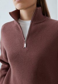 Brauner, gerippter Pullover mit hohem Kragen und halbem Reißverschluss. Weiche Textur mit metallischem Reißverschluss und darunter liegendem weißen Hemd in Lagen.
