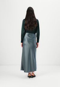 Satinová dlhá sukňa v jemnej teal farbe s priliehavým pásom a voľným strihom. V kombinácii s tmavozelenou blúzkou s dlhým rukávom. Čierne topánky na podpätku.
