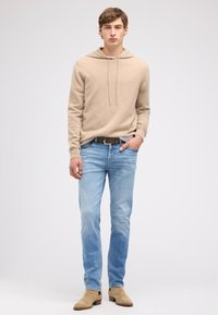 Beżowa bluza z kapturem ze sznurkami, jasnoniebieskie dżinsy z denimu oraz beżowe zamszowe botki za kostkę. Prosty design o luźnym kroju. Gładka faktura.