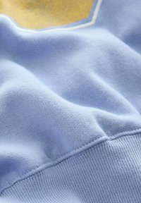 Tessuto azzurro chiaro con una texture morbida, caratterizzato da una finitura liscia e dettagli di cucitura visibili. Il design include un accento dorato.