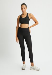Sujetador deportivo de canalé negro y leggings de talle alto, combinados con zapatillas gris claro. Textura suave, diseño ajustado y aspecto sin costuras.