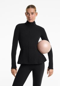 Zwarte sportjack met hoge kraag, rits aan de voorkant en uitlopende design. Model houdt een lichtroze oefenbal vast. Outfit omvat zwarte leggings.