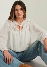 Blusa bianca con pois testurizzati, scollo arricciato, dettaglio a chiave e maniche lunghe. Abbinata a jeans azzurri chiari e stivali marroni.