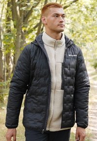 Veste extérieure matelassée noire avec une couche intérieure en polaire crème. La veste a une capuche et une fermeture éclair, avec un logo sur la poitrine.
