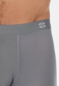 Graue Sportshorts mit glatter Textur, ausgestattet mit einem gerippten Bund und einem weißen Logo auf der Vorderseite. Flache Nähte sind sichtbar.