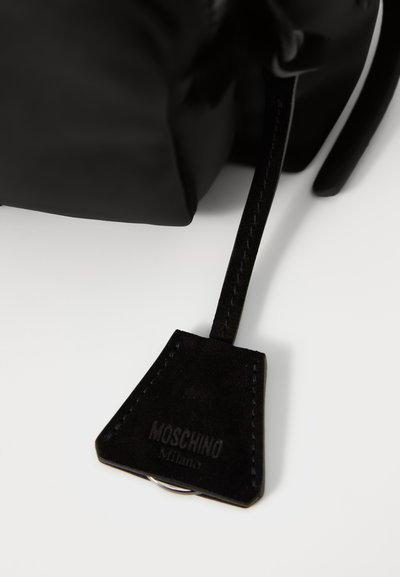 Tag de piele neagră cu o suprafață texturată, având logo-ul embosat "Moschino Milano" și un inel metalic rotund pentru atașare.