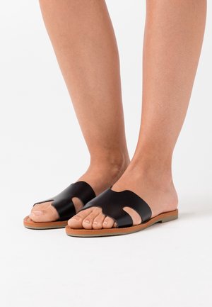 Pieds portant des sandales en cuir noires à bout ouvert avec un design découpé et des semelles plates beiges, se tenant sur une surface blanche.