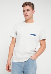 Mladý muž oblečený v svetlošedom tričku Tommy Jeans s modrým logom na vrecku a svetlomodrých džínsoch, stojaci pred jednofarebným pozadím.