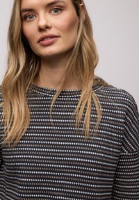 Jeune femme aux longs cheveux blonds portant un pull à motifs noir, blanc et marron, tournée légèrement vers la gauche avec un fond neutre.