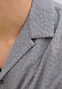 Texturirana siva košulja na dugih rukava s prepoznatljivim naboranim uzorkom, koja ima ovratnik s dijelom ovratnika vidljivim.