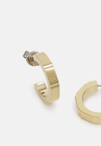 Orecchini in metallo color oro con un design testurizzato e logo "CK" inciso. Un orecchino presenta una forma a cerchio elegante; l'altro ha una forma angolare unica.