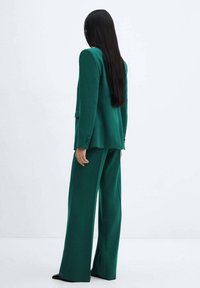 Abito verde su misura con blazer strutturato e pantaloni a gamba larga. Il tessuto appare liscio con una sottile lucentezza. Capelli lunghi neri visibili.