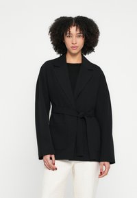 Sacou negru din material țesut, cu o curea în talie, reveruri supradimensionate și două buzunare frontale. Stilizat cu pantaloni deschiși la culoare.