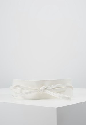 Witte leren riem met detail van een gestrikte strik, tentoongesteld op een witte sokkel tegen een effen lichtgrijze achtergrond.