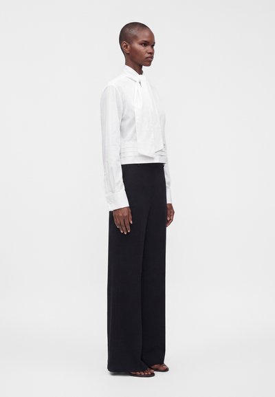 Femme debout portant un chemisier blanc à manches longues avec une cravate et un pantalon noir à jambes larges sur un fond uni.
