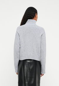 Kvinne med langt, krøllete hår iført en lys grå, spraglete turtleneck genser og et svart skinnskjørt, vendt bort fra kameraet.