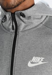 Grå zip-up hoodie i texturerat tyg med svart dragkedja, justerbara dragsnören och en vit Nike-logotyp på framsidan.