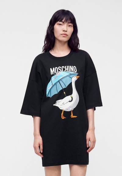 Femme portant une robe t-shirt noire oversize avec un graphisme d'une oie blanche tenant un parapluie bleu et le mot Moschino au-dessus.