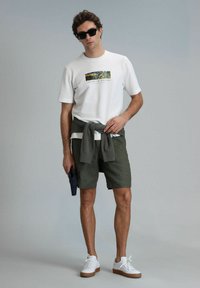 Witte t-shirt met korte mouwen met grafische print, olijfgroene shorts, grijs gebreid vest geknoopt om de taille, witte sneakers met een bruine zool.