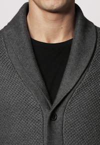 Mörkgrå sjalkrage cardigan tillverkad av strukturerat stickat tyg med en knappstängning, i kombination med en svart t-shirt under.