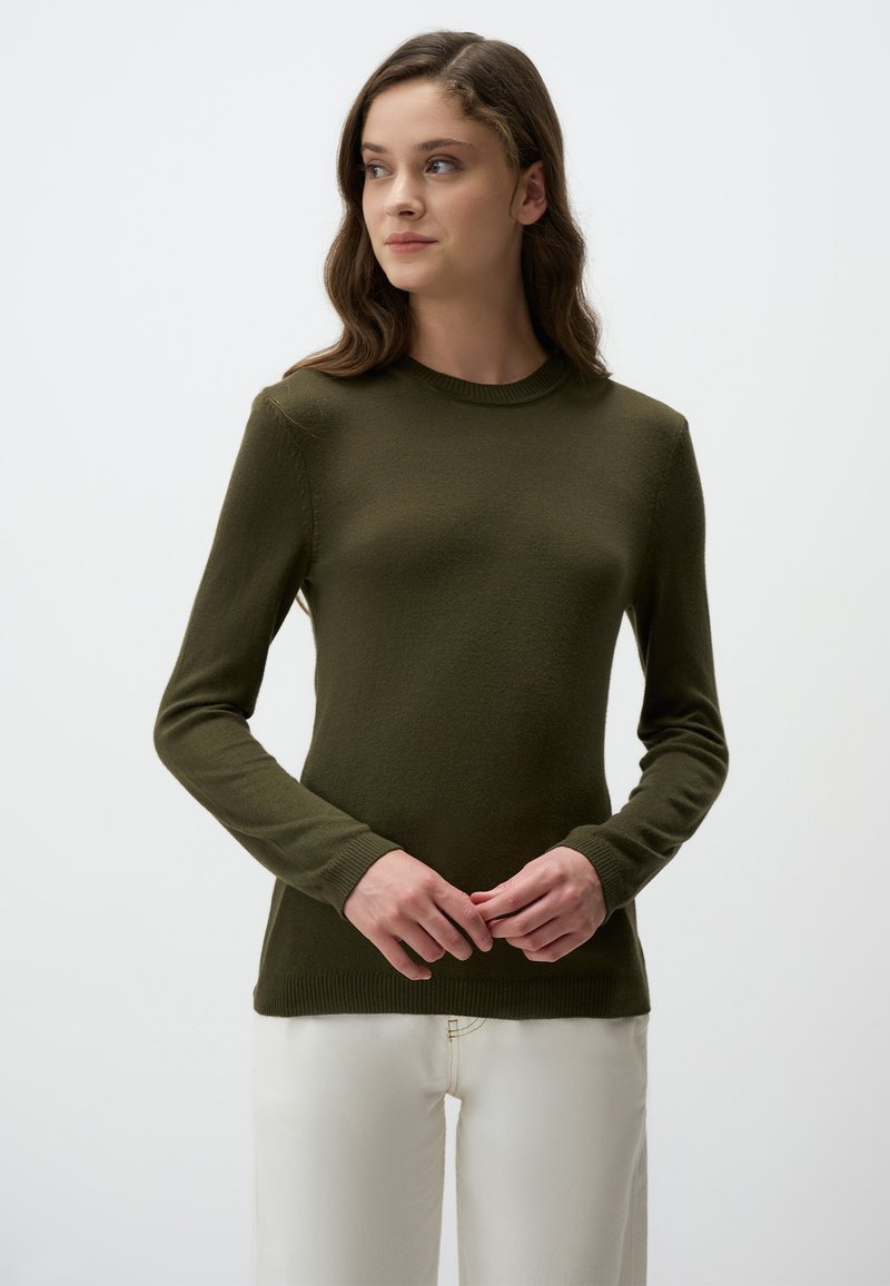 Maglione verde oliva a maniche lunghe con collo a girocollo a coste e superficie testurizzata. Indossato sopra pantaloni bianchi, evidenziando una silhouette aderente.