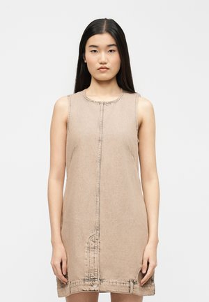 Tânără cu păr lung și închis la culoare, purtând o rochie bej din denim fără mâneci, cu cusătură frontală și detalii cu nasturi la tiv.