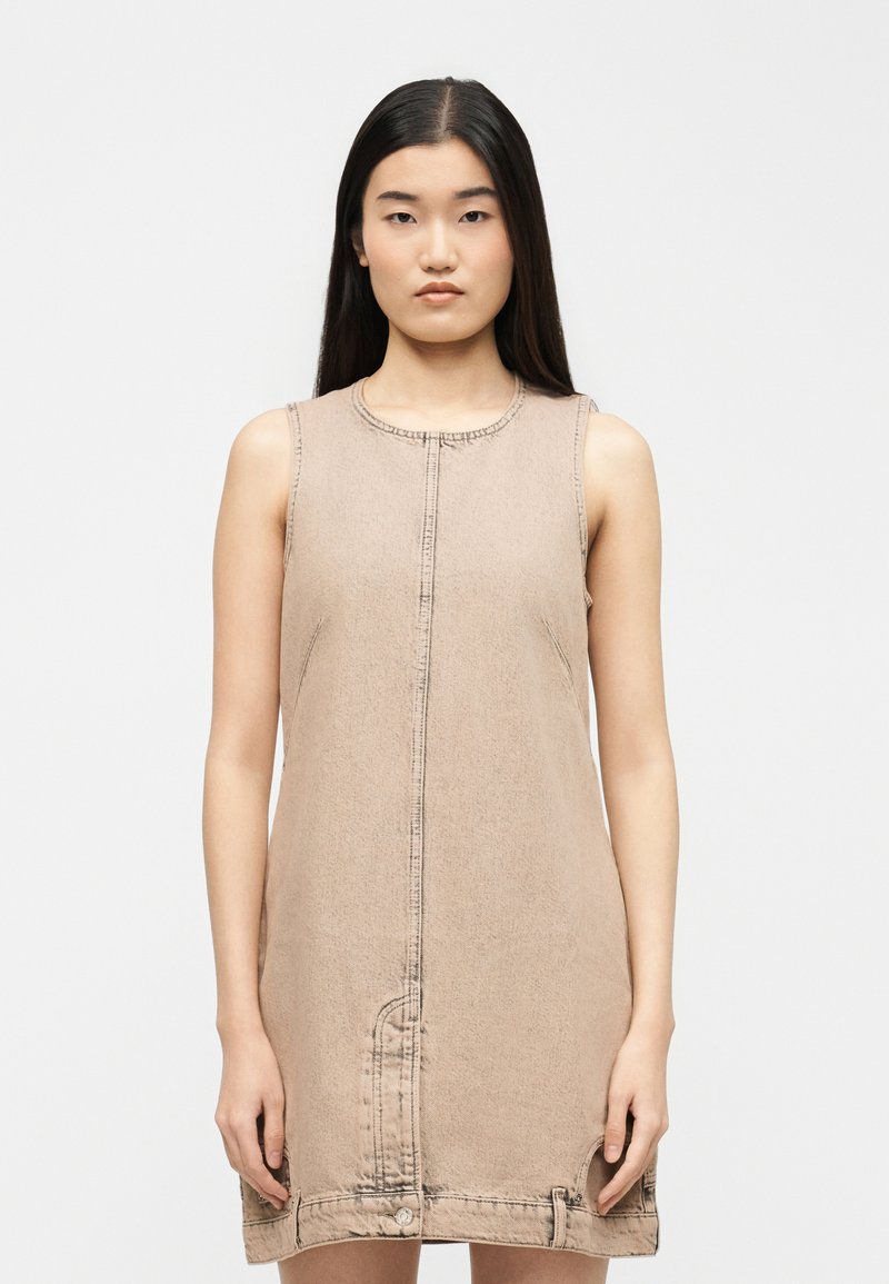Νεαρή γυναίκα με μακριά σκούρα μαλλιά που φοράει μπεζ φόρεμα από τζιν χωρίς μανίκια, με ραφή στο εμπρός μέρος και λεπτομέρειες με κουμπιά στο τελείωμα.