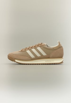 Beżowo jasno-brązowy sneaker Adidas z białym logo trzech pasków, teksturowaną tkaniną, sznurowanym zapięciem i ząbkowaną podeszwą na neutralnym tle.