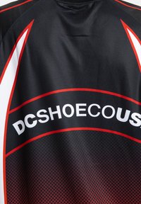 Melna sporta krekla ar sarkaniem un baltiem akcentiem, uz kura aizmugurē ar treknām baltām burtiem ir redzams zīmola nosaukums "DCSHOECOUS".