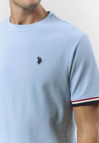 Ljusblå bomulls-t-shirt med rund hals, med en broderad logotyp på bröstet och randiga detaljer på ärmarna.