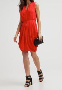 Red sleeveless dress with a wrap design, gathered waist, and flowing skirt. Paired with black strappy high-heeled sandals and a clutch.