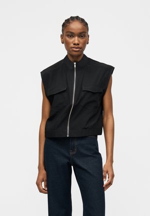 Femme aux cheveux tressés portant une veste noire sans manches zippée avec poches poitrine et un jean bleu foncé, debout devant un fond uni.