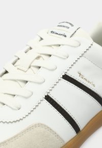 Λευκό αθλητικό παπούτσι με μπεζ σουέτ μύτη, μαύρες ρίγες στα πλευρά, λευκές κορδόνες και λογότυπο "Tamaris" στη γλώσσα και στο πλάι.