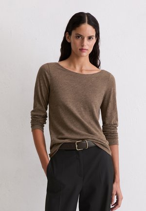 Vrouw met lang donkerhaar, gekleed in een bruin shirt met lange mouwen, ingestopt in zwarte broek met een donkere riem, staand tegen een witte muur.