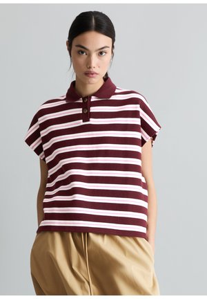 Junge Frau mit dunklen Haaren, die ein kurzärmeliges, bordeauxrotes und weiß gestreiftes Poloshirt sowie beige Hosen vor einem schlichten Hintergrund trägt.