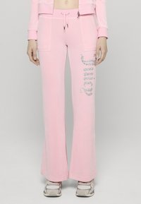 Pink bővülő szárú melegítőnadrág puha anyagból, elöl két zsebbel és ezüst csillámfeliratokkal az oldalon. Fehér sportcipővel viselve.