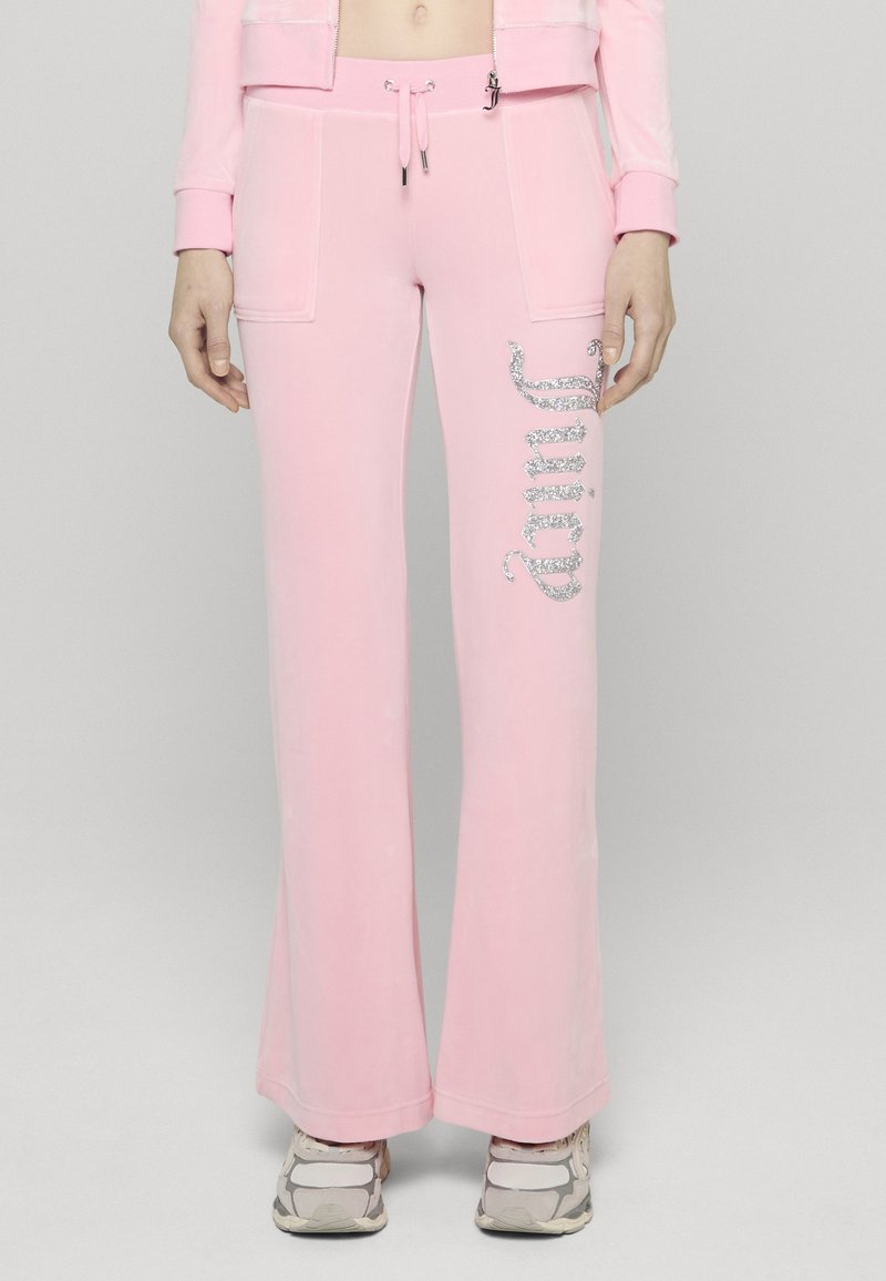 Pink bővülő szárú melegítőnadrág puha anyagból, elöl két zsebbel és ezüst csillámfeliratokkal az oldalon. Fehér sportcipővel viselve.