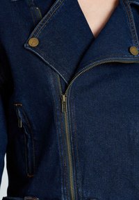 Chaqueta de mezclilla azul oscuro con cierre de cremallera en el frente, solapas anguladas y acentos de hardware en latón. Presenta detalles de costura y dos bolsillos delanteros.