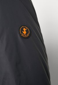 Schwarze Stoffjacke mit einem runden orangen „Save the Duck“-Logo-Patch auf der linken Seite. Das Material wirkt glatt und leicht.