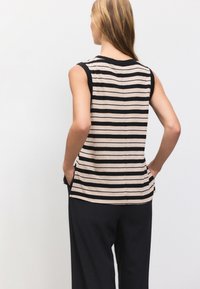 Brezrokavnik z beločrnimi črtami v bež in črni barvi. Ima okrogel izrez, stranske žepe in sproščen kroj. Oblečen s temnimi hlačami širokih nog.