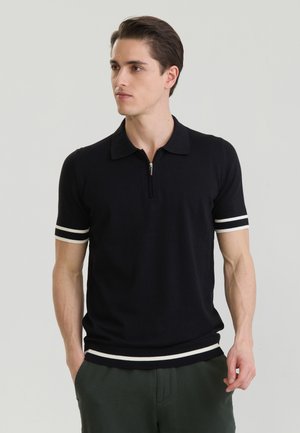 Homme portant un polo noir à manches courtes avec des rayures blanches sur les manches et l'ourlet, debout les mains dans les poches, regardant vers la gauche.