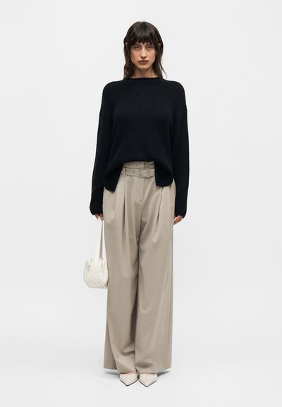 Γυναίκα με μαύρο πλεκτό πουλόβερ, μπεζ παντελόνια με φαρδιά πόδια και ζώνη, λευκά μυτερά παπούτσια και κρατά μια μικρή λευκή πλεκτή τσάντα.