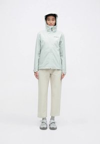 Světle zelená nepromokavá bunda s kapucí, předním zipem a logem. Kombinována s krémovobílými volnými kalhotami a šedou obuví.