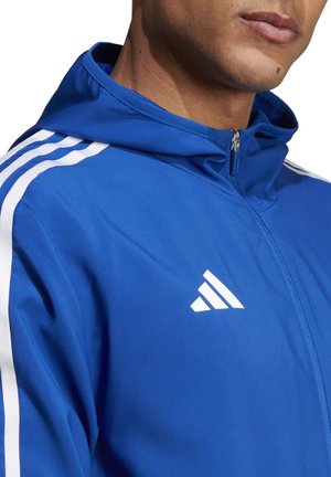 Blauwe sportjack met witte strepen langs de schouders, ritsluiting en klein wit logo. Glad textuur, capuchon ontwerp.