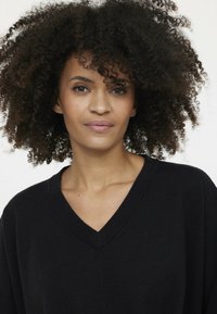 Schwarzer V-Ausschnitt-Pullover mit lockerer Passform und geripptem Ausschnitt. Das Modell hat lockiges, schulterlanges Haar und einen neutralen Hintergrund.