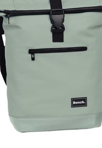 Sac à dos étanche vert avec un design à rabat, quincaillerie noire, poche zippée à l'avant et étiquette de logo en bas à l'avant. Texture mate.