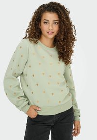 Sweatshirt vert clair avec un col ras du cou et des poignets côtelés, présentant des motifs de cœurs dorés sur l'ensemble, associé à un jean foncé.