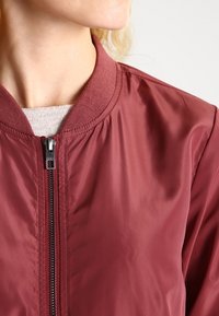 Bordová bomber bunda s hladkým, lesklým povrchem, žebrovaným límcem a středovým zipem. Minimalistický design bez viditelných vzorů nebo akcentů.